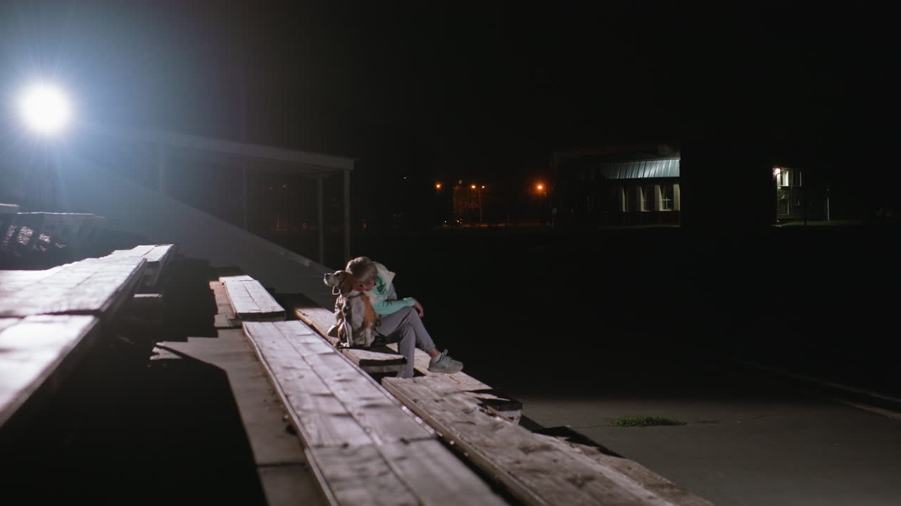 Canine behaviourist seated peacefully on platform with german shepherd at night, resting her head gently on dog while crossing her legs, surrounded by empty benches under strong spotlight
