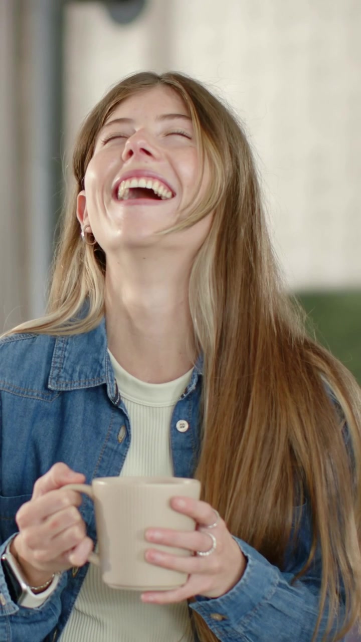 Video verticale: Reazione a una battuta, donna in camicia di jeans che tiene una tazza beige e ride in un caffè.