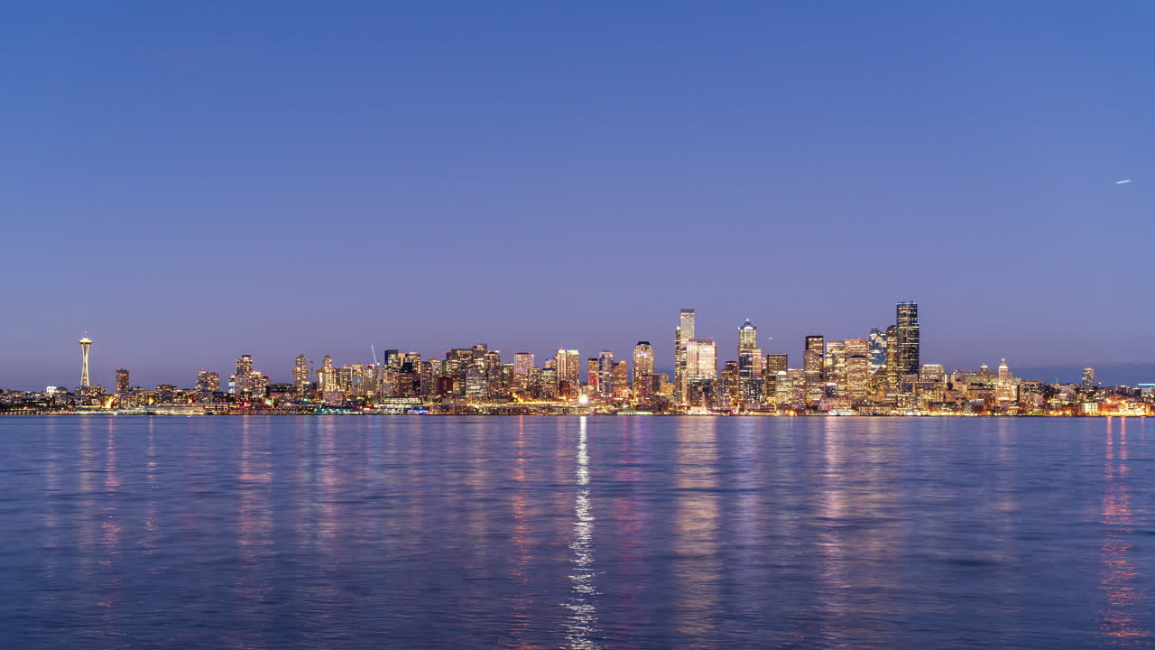 A timelapse video capturing the glowing Seattle skyline at night, with city lights reflecting off the calm waters of Puget Sound under a clear sky.