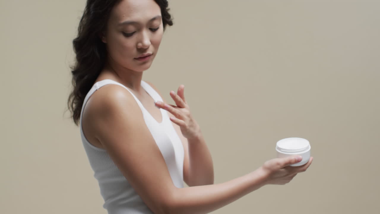 어두운 머리카락의 아시아 여성이 베이지색 배경에 피부에 크림을 바르며, 복사 공간, 느린 동작