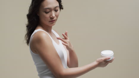 mujer asiática con cabello oscuro aplicando crema en su piel sobre un fondo beige, espacio de copia, cámara lenta