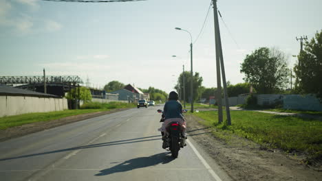 two ladies ride on a power bike at a slow pace along a calm road, two cars approach from the opposite direction, electric poles, trees, and buildings are visible in the background