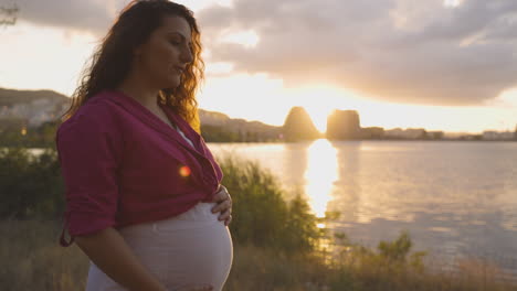 mamá embarazada pensativa al atardecer sonriendo y acariciando el vientre frente al mar