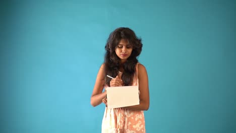 A-young-Indian-girl-in-orange-frock-writing-in-a-notepad-and-pencil-standing-in-an-isolated-blue-background