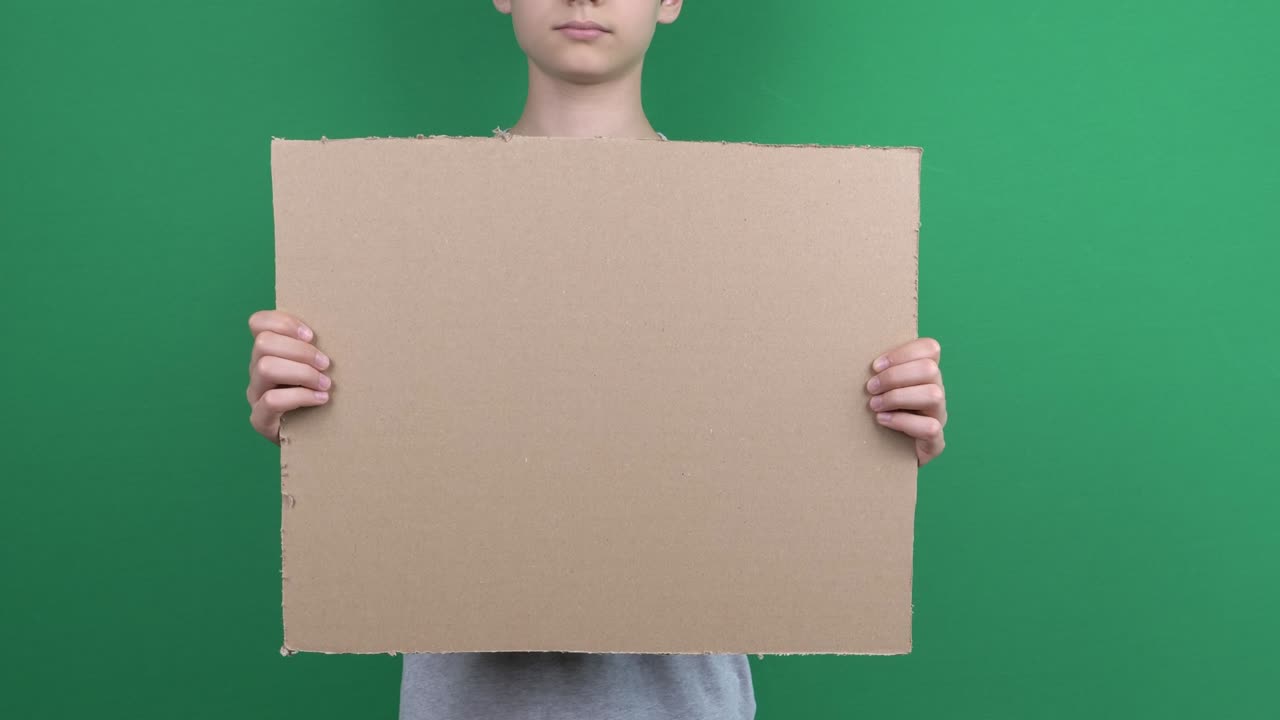 Boy holding in his hands blank empty cardboard poster on green chromakey screen background