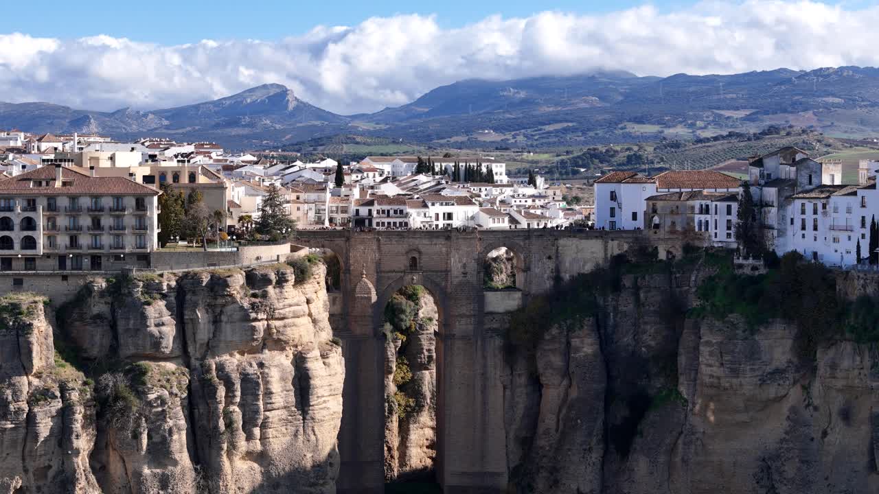 Ronda, Spain, a breathtaking panning aerial view capturing the iconic Puente Nuevo bridge, dramatic cliffs, charming white houses, and a picturesque cloudy sky creating a captivating landscape