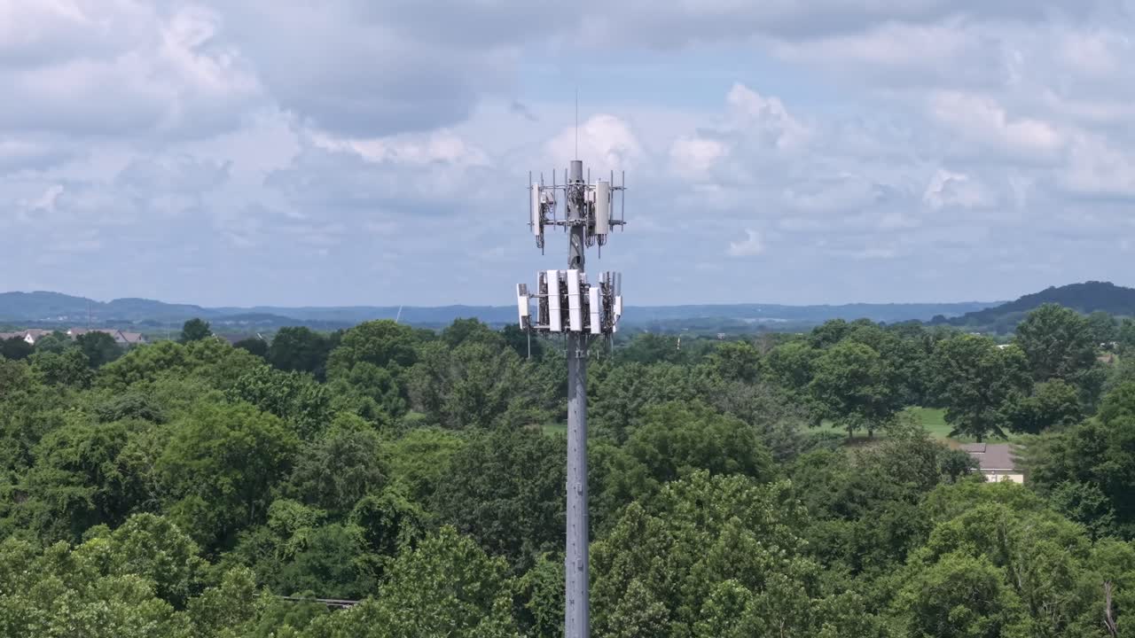 Cell tower equipment providing network signal above large area of trees, aerial obit with cloudy blue sky