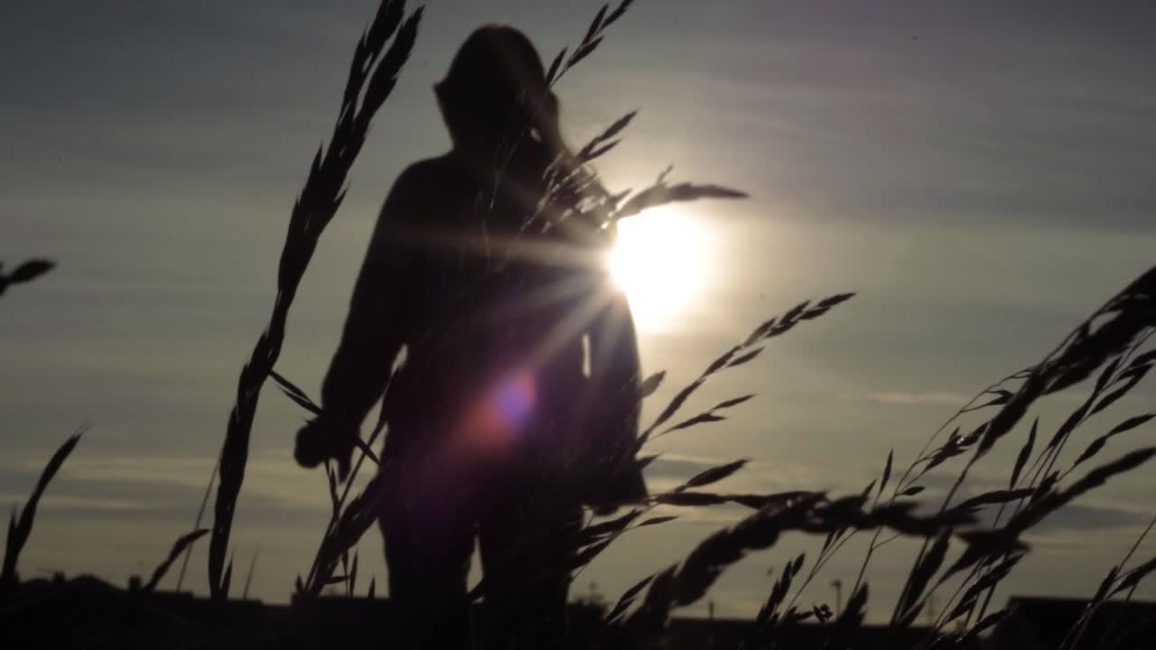 Silhouette of woman walking through tall grass against sky at sunset medium shot