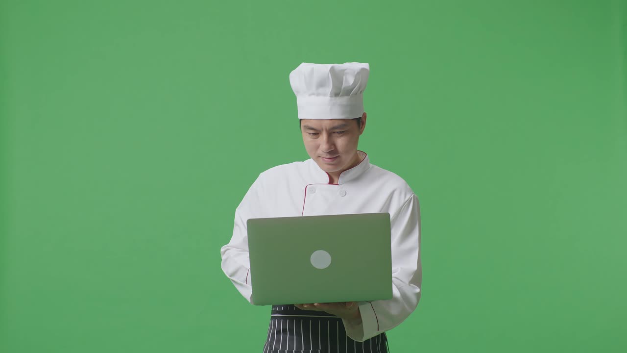 Asian Man Chef Smiling And Using A Laptop While Standing In The Green Screen Background Studio
