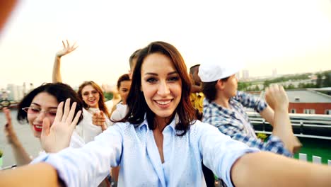 point of view shot of young woman holding camera and recording rooftop party with happy people dancing, laughing and looking at camera. youth and fun concept.