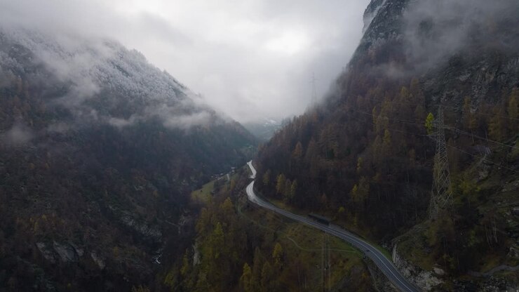 Moody backward flying drone aerial shot through Swiss alps forest mountain road with clouds covering steep mountain side and snow covered trees on cloudy winter afternoon with car driving on road