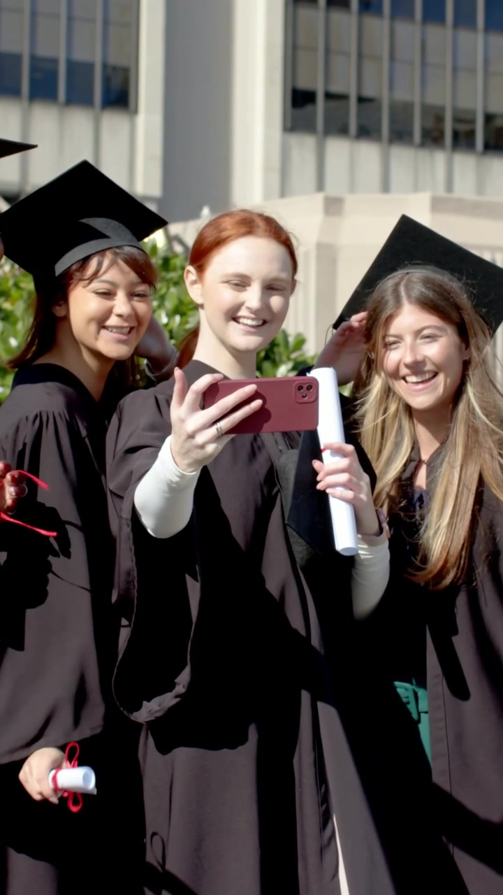 Video verticale: Alzando il telefono, 3 laureati in toga che si scattano selfie all'università, tenendo i diplomi e festeggiando.