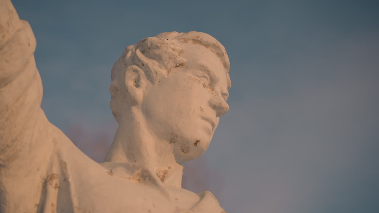 Close up of classical sculpture captured under bright sky with soft light revealing weathered surface texture, while distant bare tree appears blurred in peaceful background