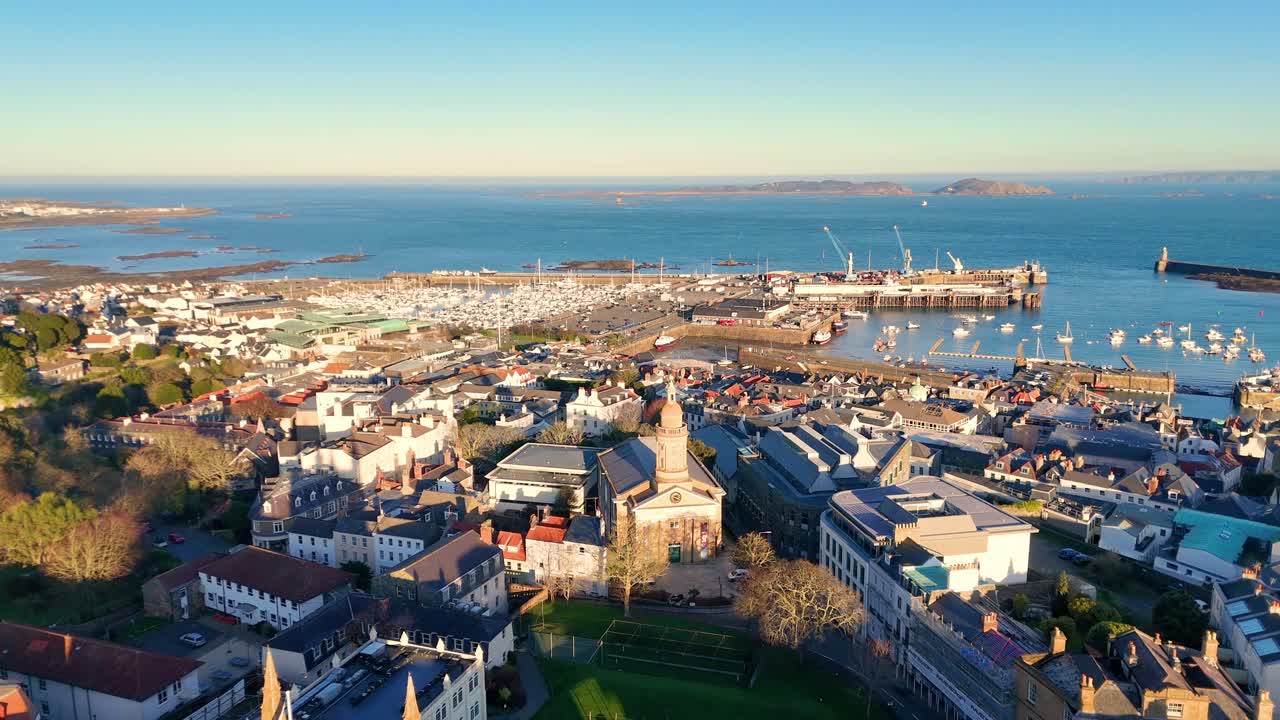 High reveal drone footage of the roof tops of St Peter Port Guernsey in the golden hour featuring Elizabeth College with views across town and the harbour to Herm and Jethou