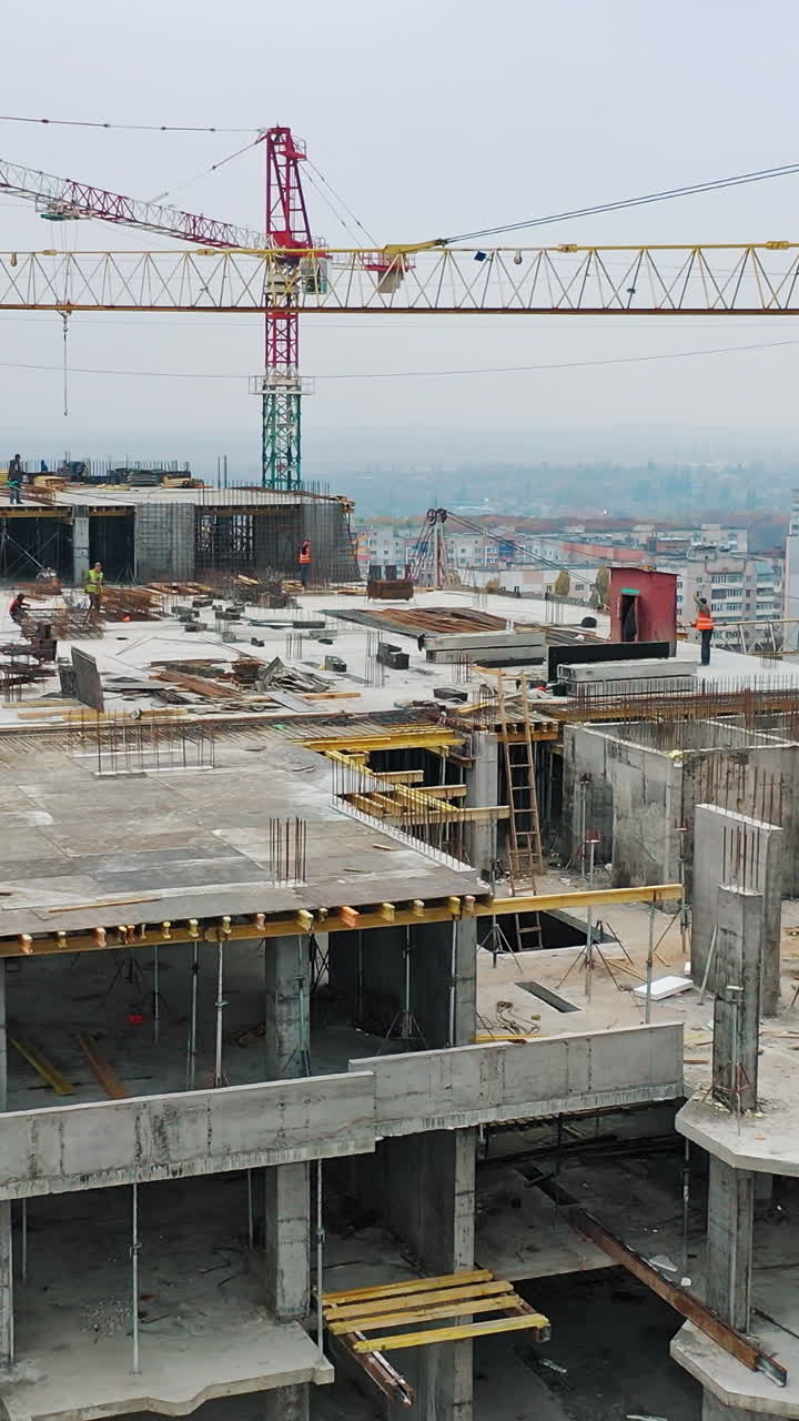 New construction site of shopping center. Aerial view on roof with workers and big cranes during construction on the city background. Drone moves back. Vertical video