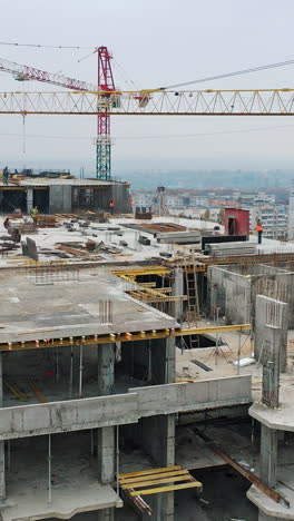 new construction site of shopping center. aerial view on roof with workers and big cranes during construction on the city background. drone moves back. vertical video