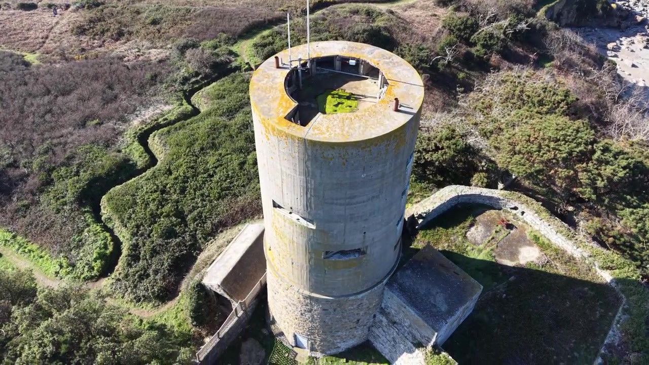 L’Eree Guernsey: Dramatische Drohnenaufnahme eines alten deutschen Wachturms auf der Landzunge, der sich vor strahlend blauem Himmel nähert und direkt über dem Turm mit einer Detailansicht in der späten Nachmittagssonne endet