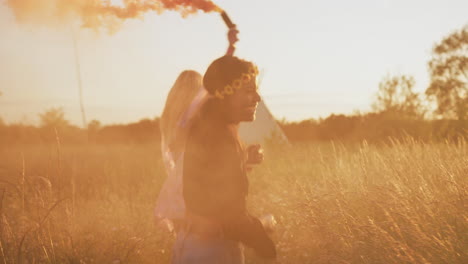 Two-Female-Friends-Camping-At-Music-Festival-Running-Through-Field-With-Smoke-Flares