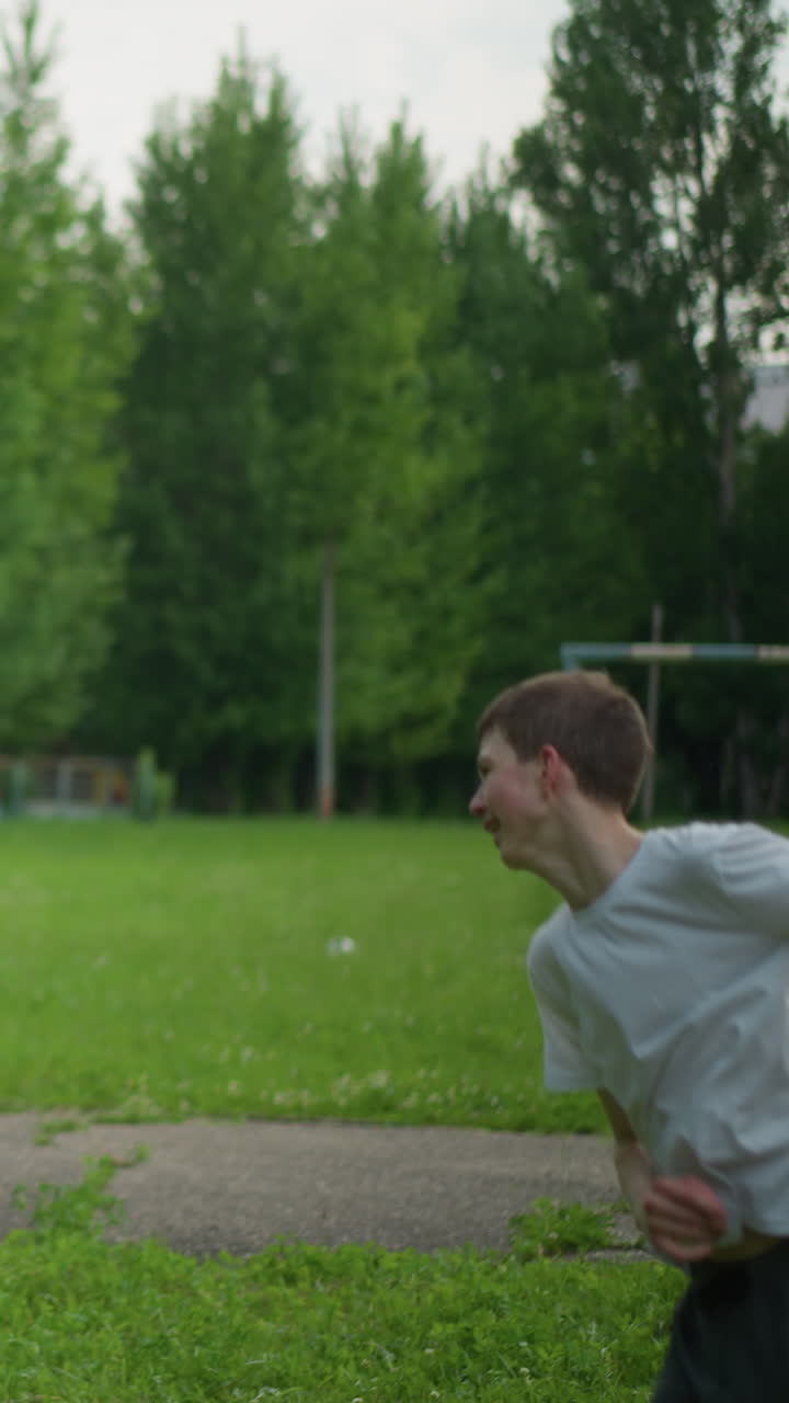 A father throws a soccer ball to his son, who skillfully heads it away in a grassy field with a blurred view of a paved road and greenery in the background
