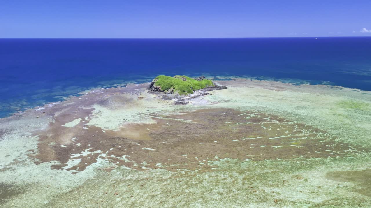 Cinematische drone-nadering over een ondiepe, levendige koraalriflagune richting een klein rotsachtig eilandje onder een helderblauwe lucht in Ishigaki, Okinawa.