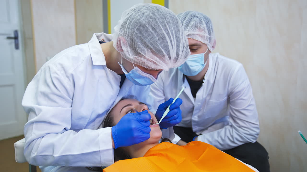 Future dentists sitting bent over the girl. One student checks lower row of teeth with medical instruments and another watches him.