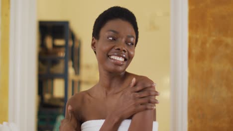 Portrait-of-smiling-african-american-woman-with-towel-touching-her-skin-in-bathroom