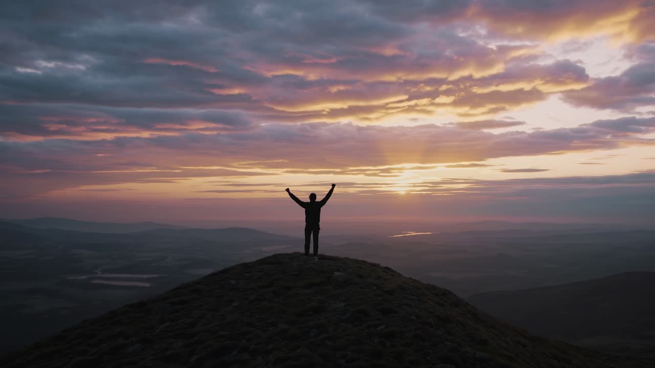 Silueta de una persona en la cima de una colina al atardecer, con los brazos levantados. Este vídeo se ha grabado desde un ángulo bajo.