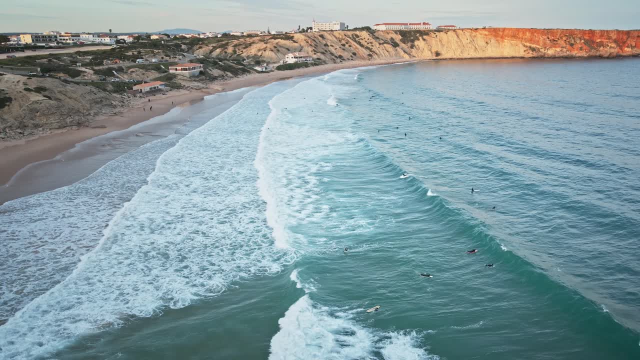 High altitude drone shot looking straight down at surfers riding waves in turquoise Atlantic water near the sandy shore of Sagres, Portugal