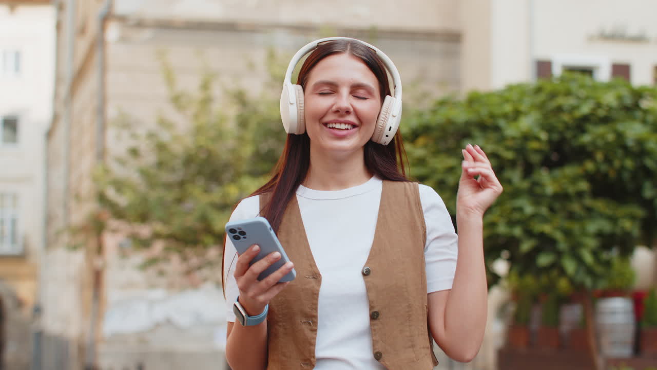 mujer joven encantada con auriculares inalámbricos eligiendo escuchar música sosteniendo un teléfono inteligente en la calle