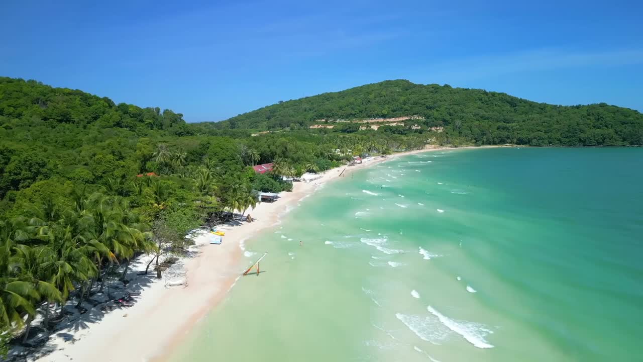 Aerial view of a tropical beach with turquoise waters, white sand, and lush green hills under a clear blue sky.