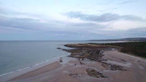 imágenes de drones en movimiento lento que se elevan por encima de una playa de arena y hacia la luna creciente al atardecer mientras la marea golpea la orilla