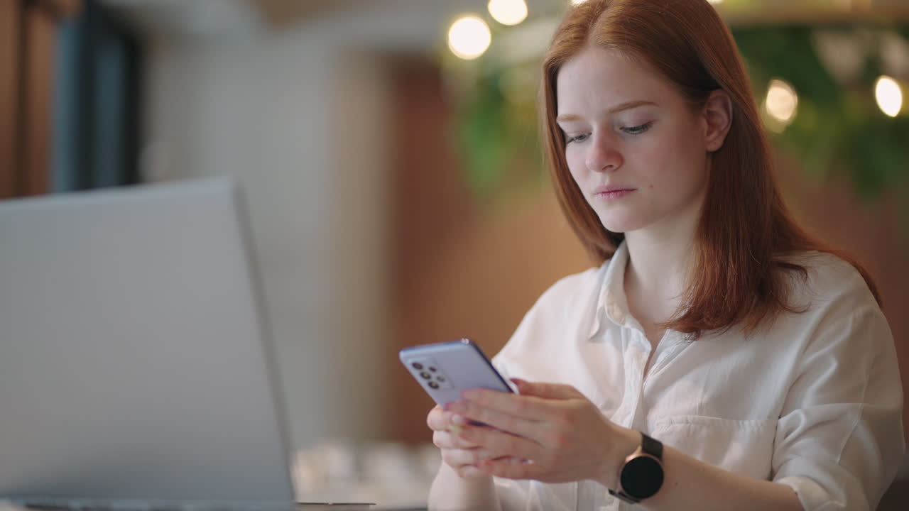 Red-haired Woman working from home using laptop computer while reading text message on mobile phone. Woman using a phone. Serious charming woman using smartphone while working with laptop at home