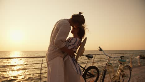 un tipo con ropa ligera y una chica con una camisa blanca azul están abrazándose en una playa que está cubierta de tablas cerca de sus bicicletas cerca del mar al amanecer en verano