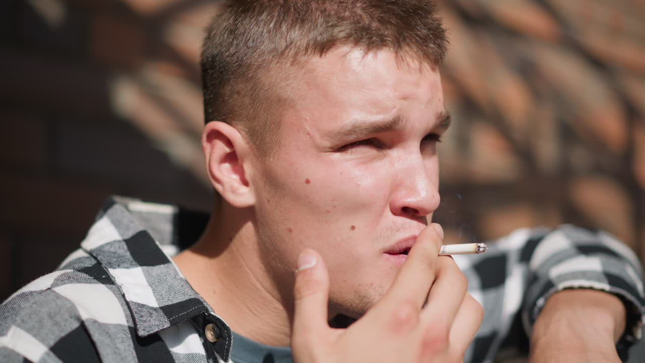 close up of young man with spotty face smoking cigarette while resting on iron railing in bright sunlight with soft blur background and dramatic shadow play across brick wall behind