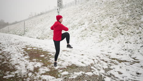 mujer realizando ejercicios dinámicos de invierno al aire libre en terreno nevado, con capucha roja y guantes negros, rodeada de colina nevada con rieles de hierro y atmósfera de niebla