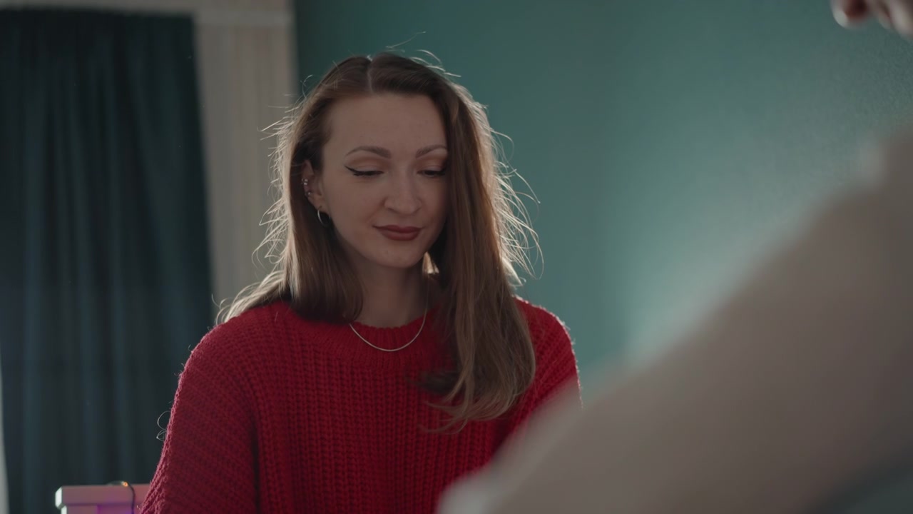 Young woman in red sweater looks down with calm smile while foreground hand adjusts object along rail beside teal curtain, creating quiet indoor home moment with soft daylight and gentle motion