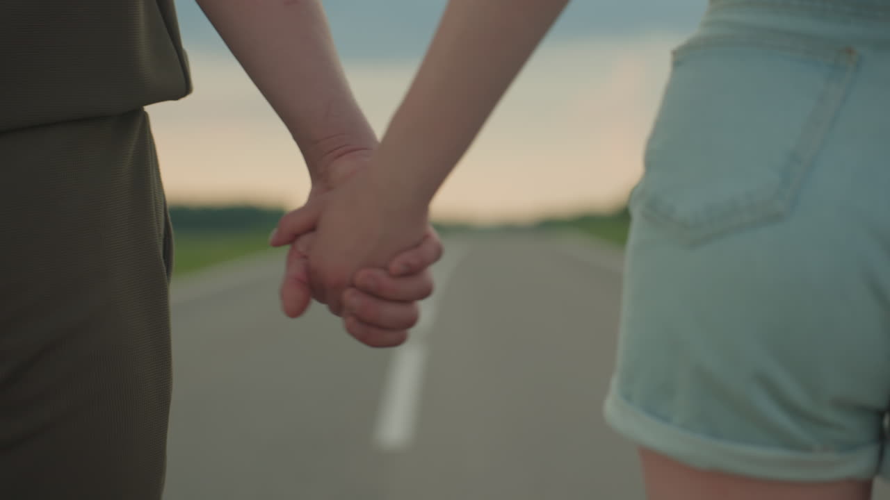 close up rear view of couple holding hands while walking empty country road under sunset sky conveying romance travel journey in casual summer clothes by green fields