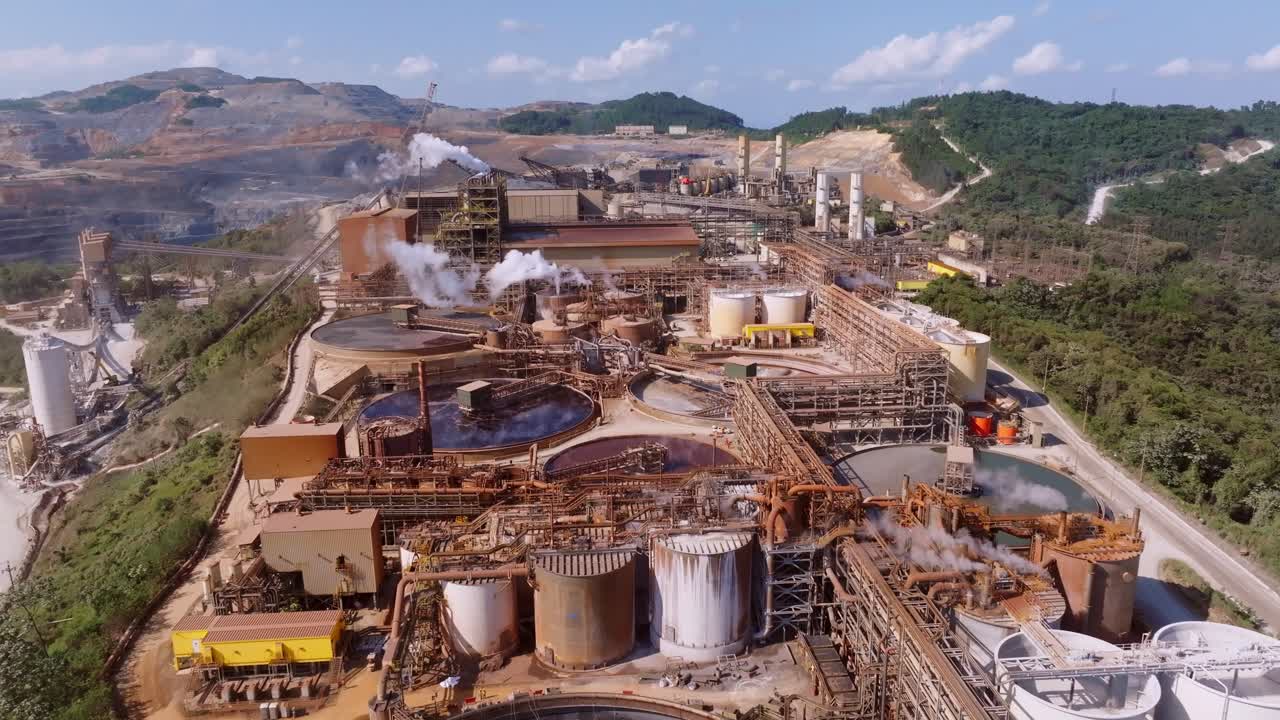 Slow drone flight over industrial mine factory in Dominican Republic. Aerial flyover. Ponds and pipelines with rising steam of smokestack. Wide shot.
