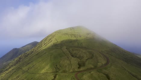 drone orbiting vulcanic mountain, pico da esperança, covered in lush green with low clouds in são jorge island, the azores, portugal