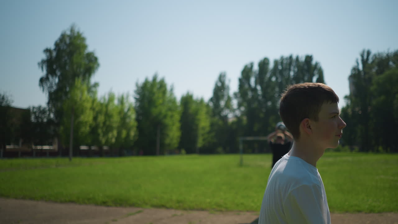 A young boy heads a soccer ball outdoors, while in the blurred background, another person is holding a ball up