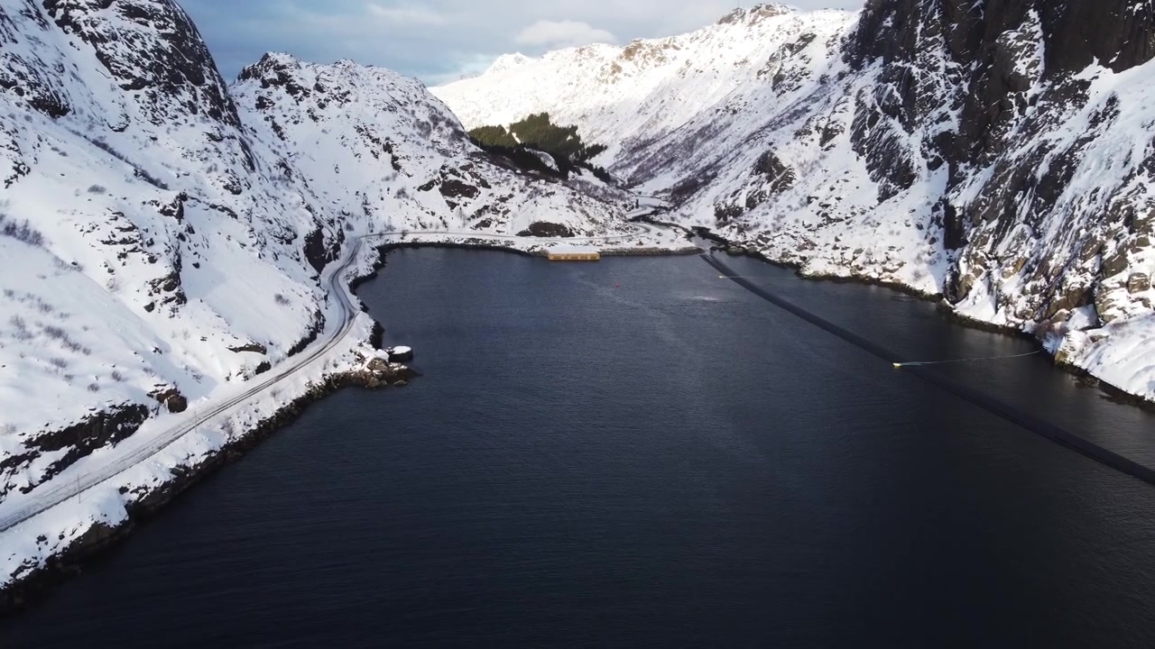 Dronebilde som flyr over det mørke vannet i en smal fjord på Lofoten, Norge, omgitt av bratte, snødekte fjellrygger under den arktiske vintersesongen.