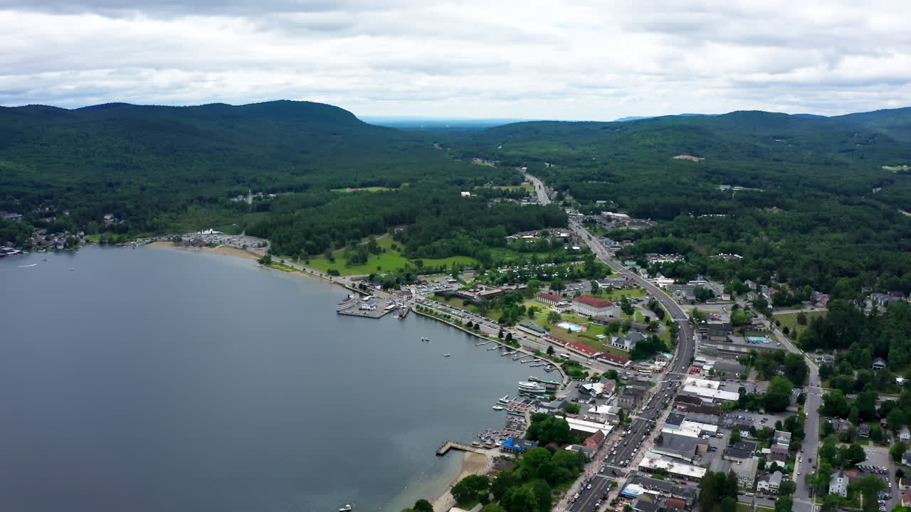 Drone fly in toward Lake George Village in New York, revealing marinas, lakeside streets, beaches, and the surrounding Adirondack Mountains stretching beyond the waterfront town