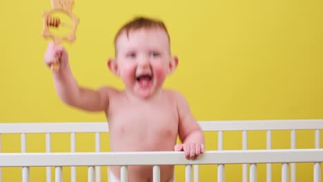 happy toddler baby boy in a crib with a wooden toy in his hand, yellow studio background. a smiling child in white diaper