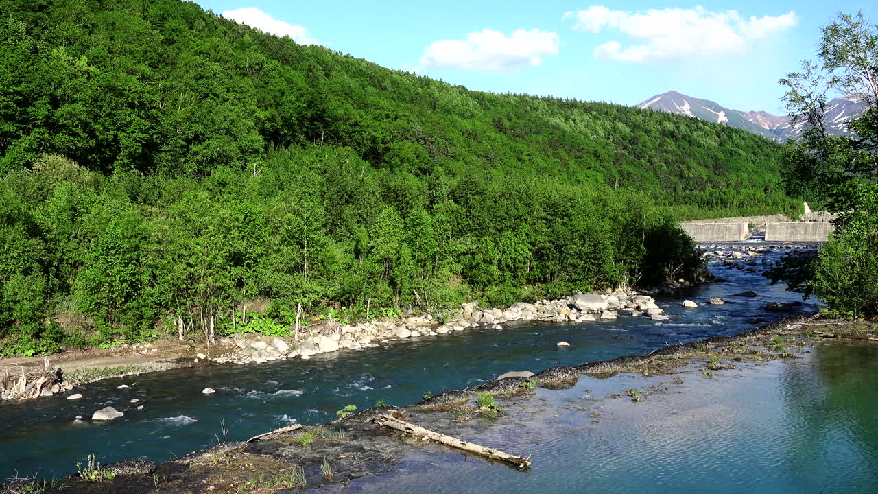 Blue Pond relaxing river flowing its water through rocks next to an imposing mountain of abundant green vegetation under a beautiful clear sky in a wonderful landscape in Hokkaido Shirogane Japan