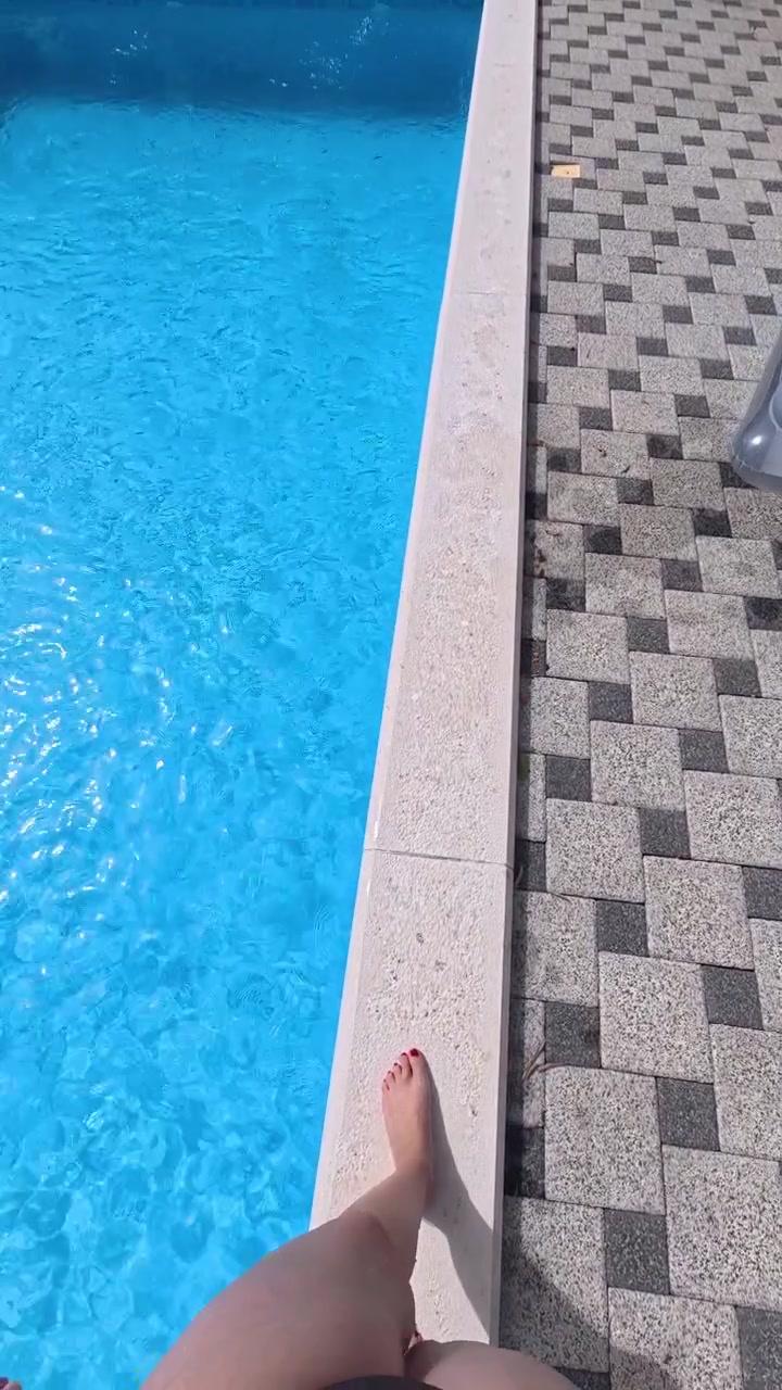 First person view of a woman walking along the edge of a clear blue outdoor swimming pool, capturing a casual summer leisure and vacation moment.
