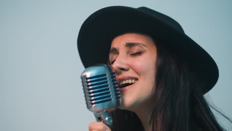 cantante femminile appassionata con cappello nero e giacca di pelle che canta in un microfono vintage, muovendosi espressivamente contro uno sfondo bianco morbido, vista ravvicinata con zoom della telecamera