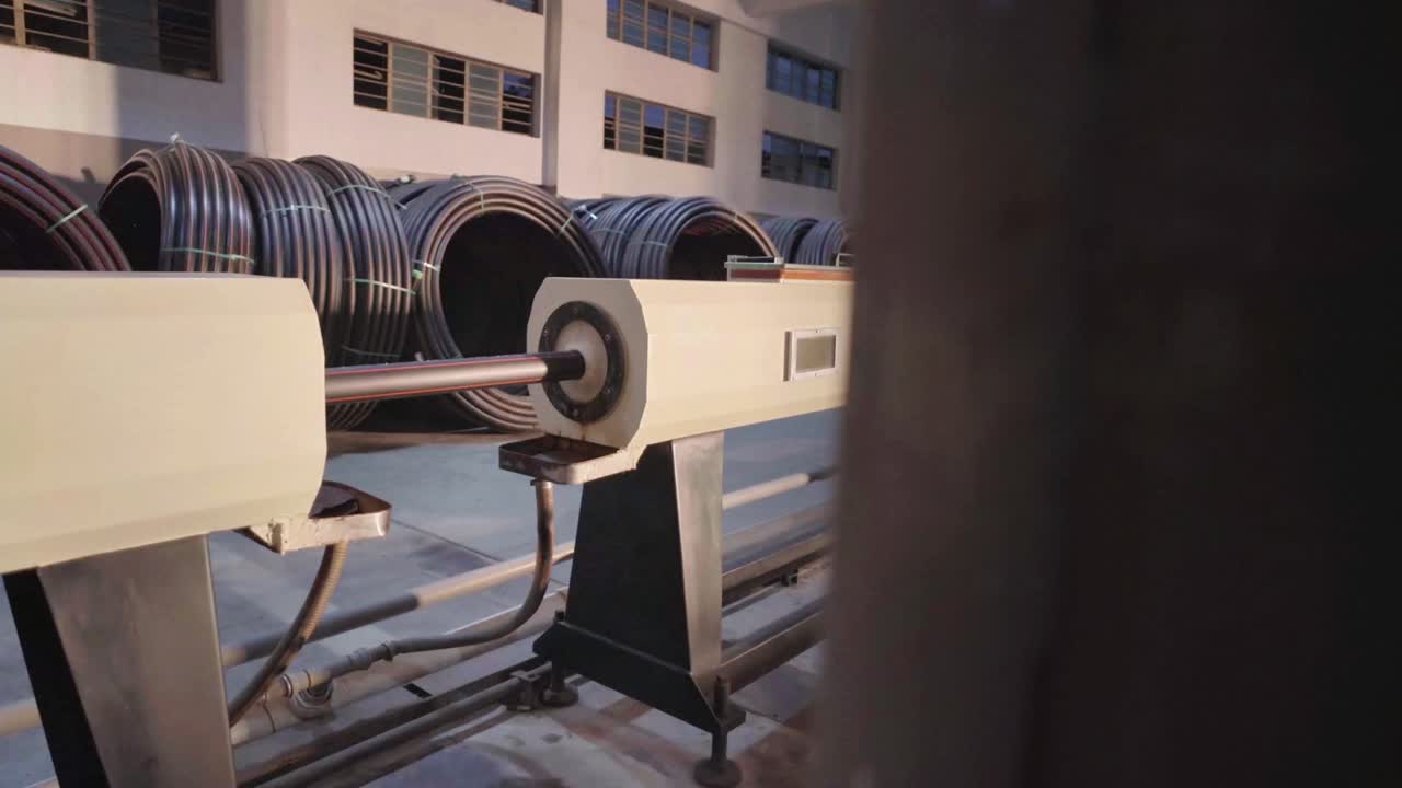 Interior of a factory featuring long metallic pipes and machinery arranged in a line. The scene captures the industrial environment and manufacturing setup.