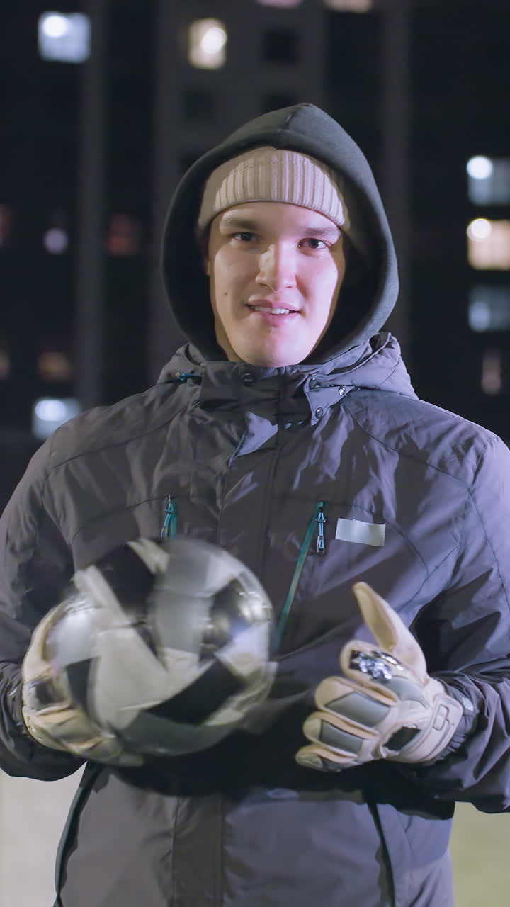 atleta sonriente con capucha y guantes alterna la pelota de fútbol en las manos y da pulgares hacia arriba con la mano izquierda durante el entrenamiento nocturno en el campo deportivo urbano rodeado de edificios iluminados de la ciudad