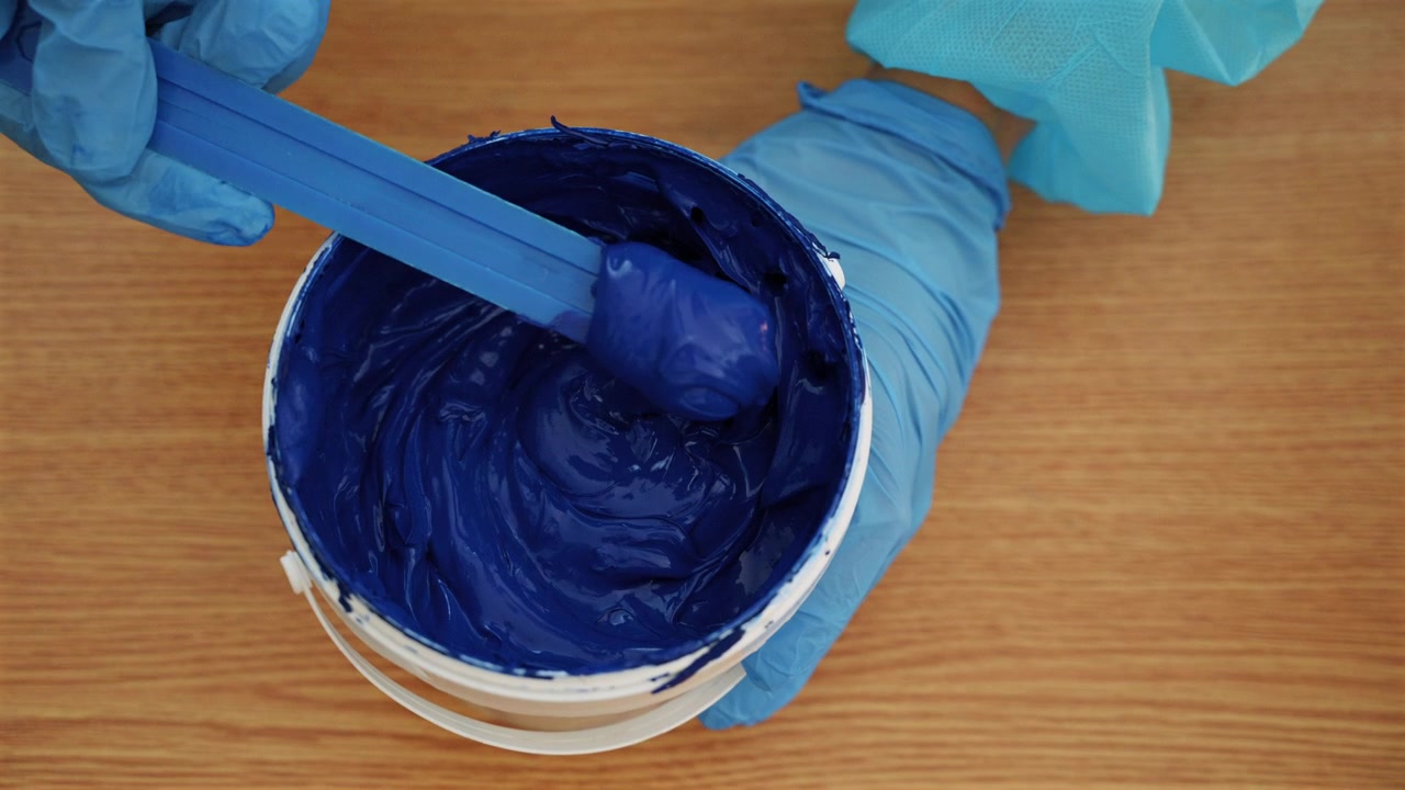 Close-up de tinta azul espessa para serigrafia sendo mexida em um recipiente por um trabalhador com luvas.