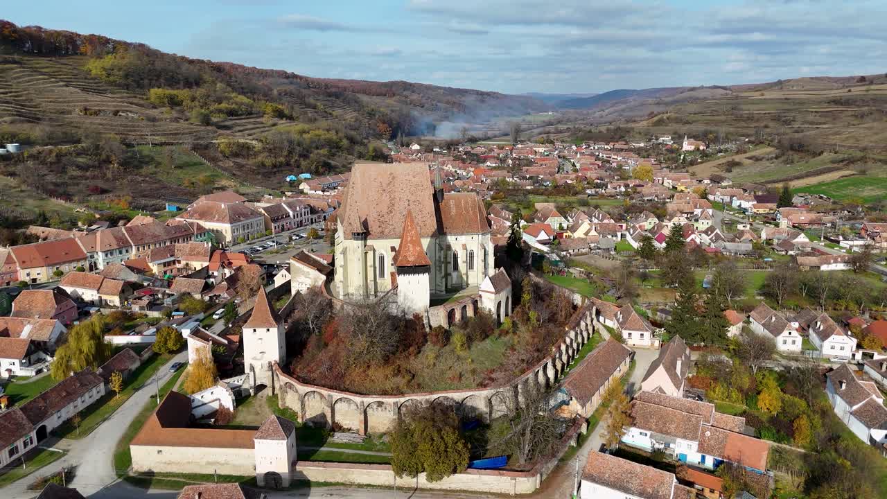 As the drone moves closer, the vibrant autumn colors of the surrounding landscape and the charming village below come into view, highlighting the seamless blend of rich history, medieval architecture